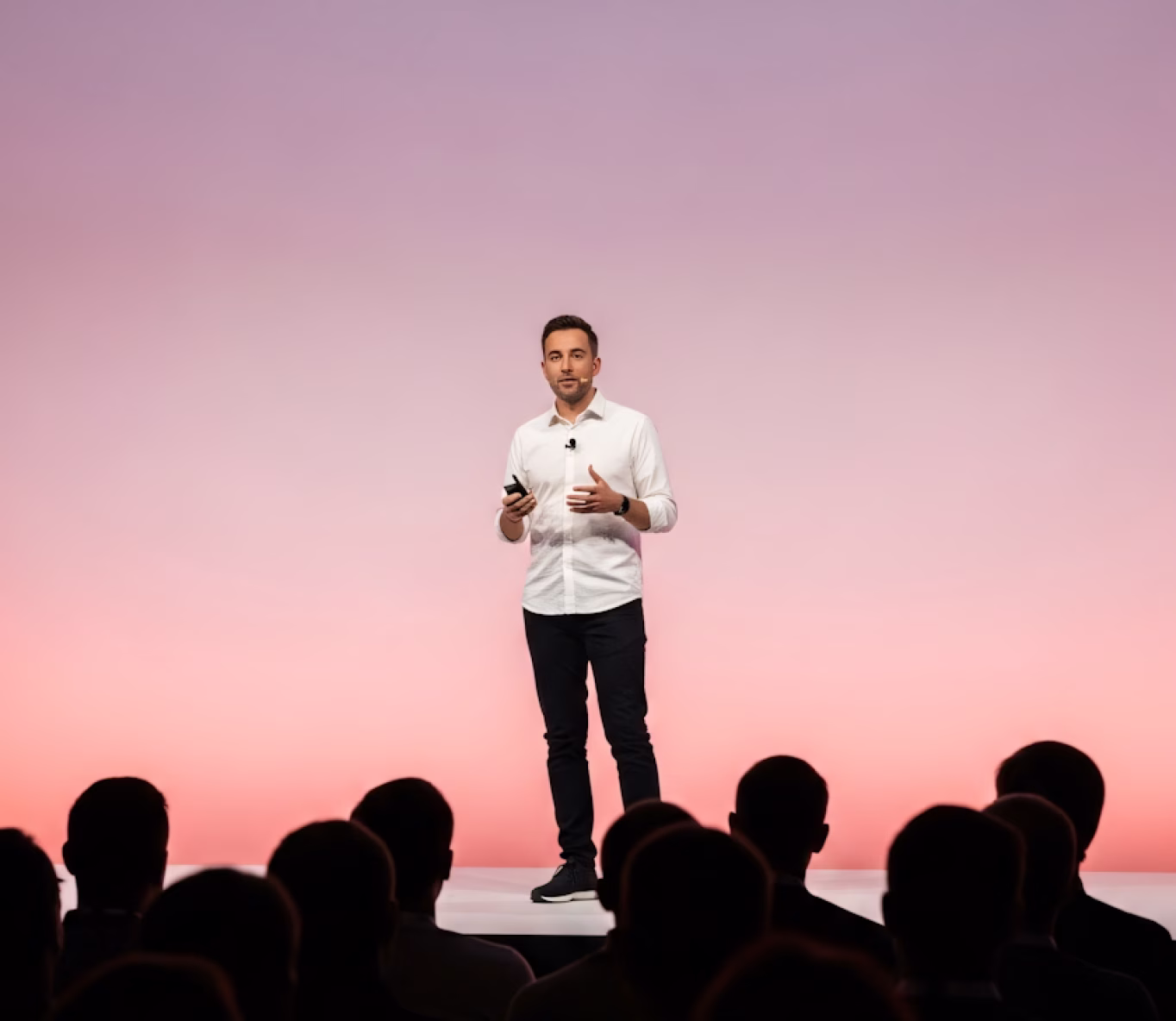 Man in white shirt and black pants giving a presentation on stage with a pink gradient background and audience silhouettes in foreground.